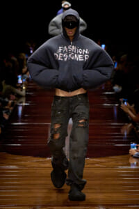 Model walks a fashion show in an oversized navy hoodie with 'FASHION DESIGNER' text, cropped midriff, and ripped black jeans on a glossy wooden runway.
