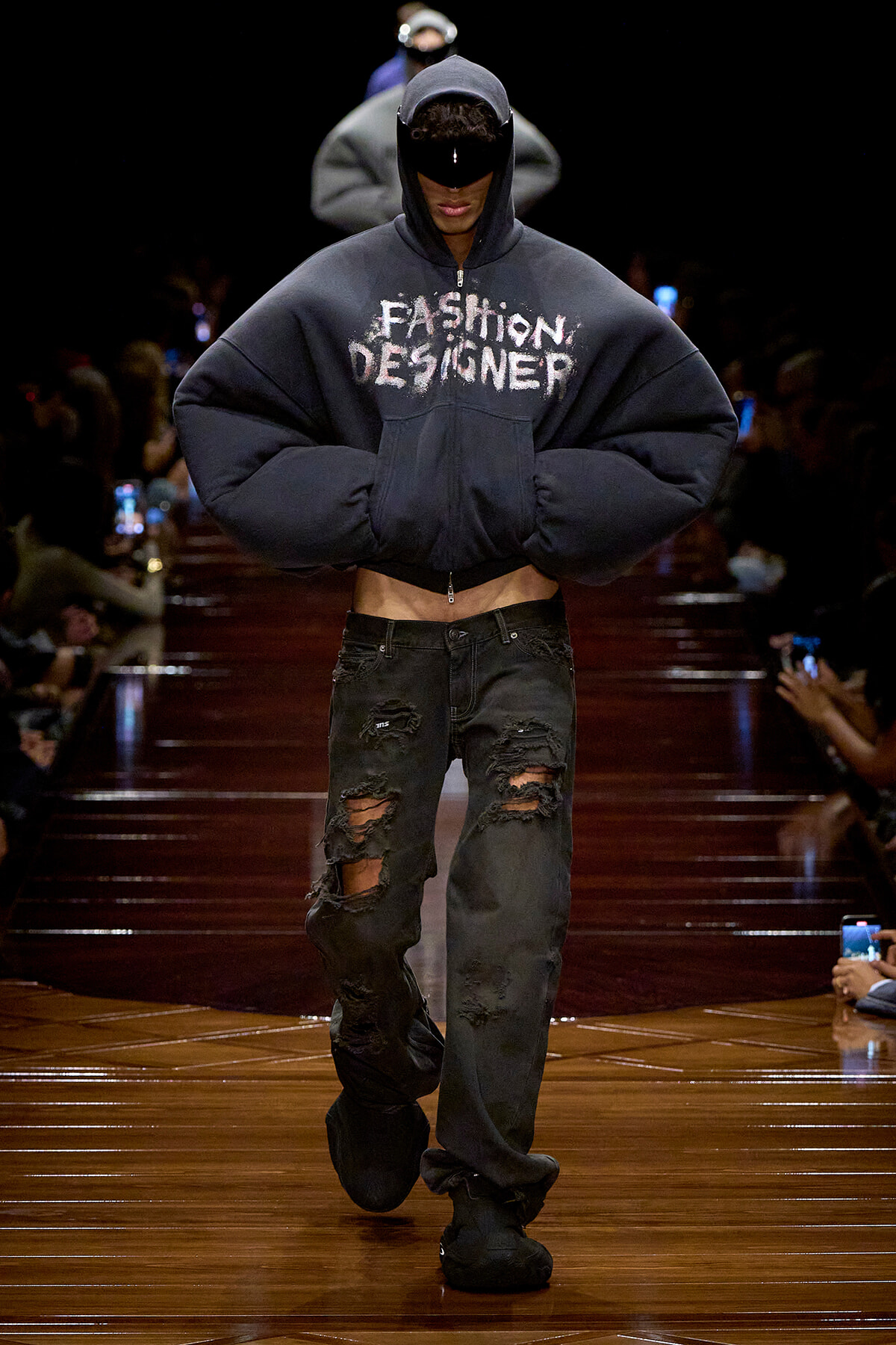 Model walks a fashion show in an oversized navy hoodie with 'FASHION DESIGNER' text, cropped midriff, and ripped black jeans on a glossy wooden runway.