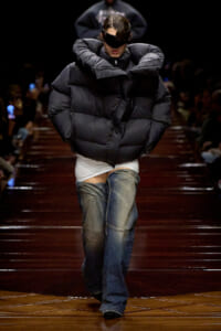 Model on a runway wearing an oversized black puffer jacket, white top, and faded jeans on a glossy wooden catwalk.