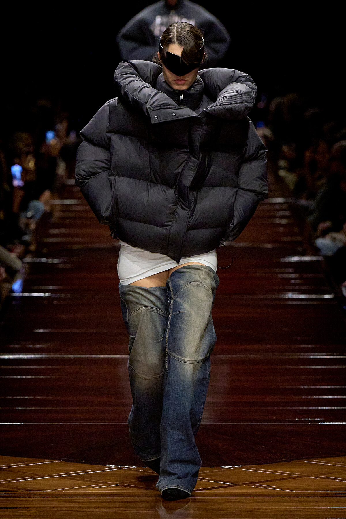 Model on a runway wearing an oversized black puffer jacket, white top, and faded jeans on a glossy wooden catwalk.