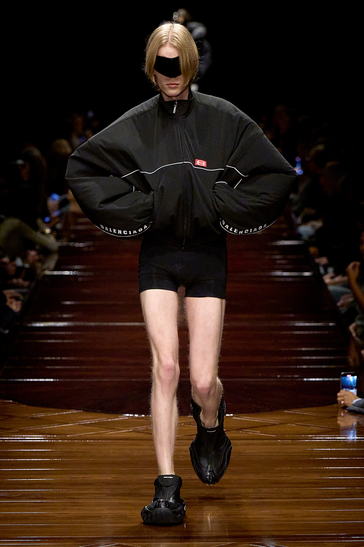 Runway model wearing an oversized black jacket with white piping, black shorts, and a black eye mask, walking on a glossy wooden catwalk with Balenciaga branding on sleeves.