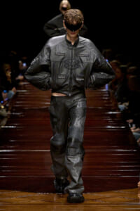 Male model on a dark runway wearing a black leather Balenciaga outfit with a blindfold, hands on hips.