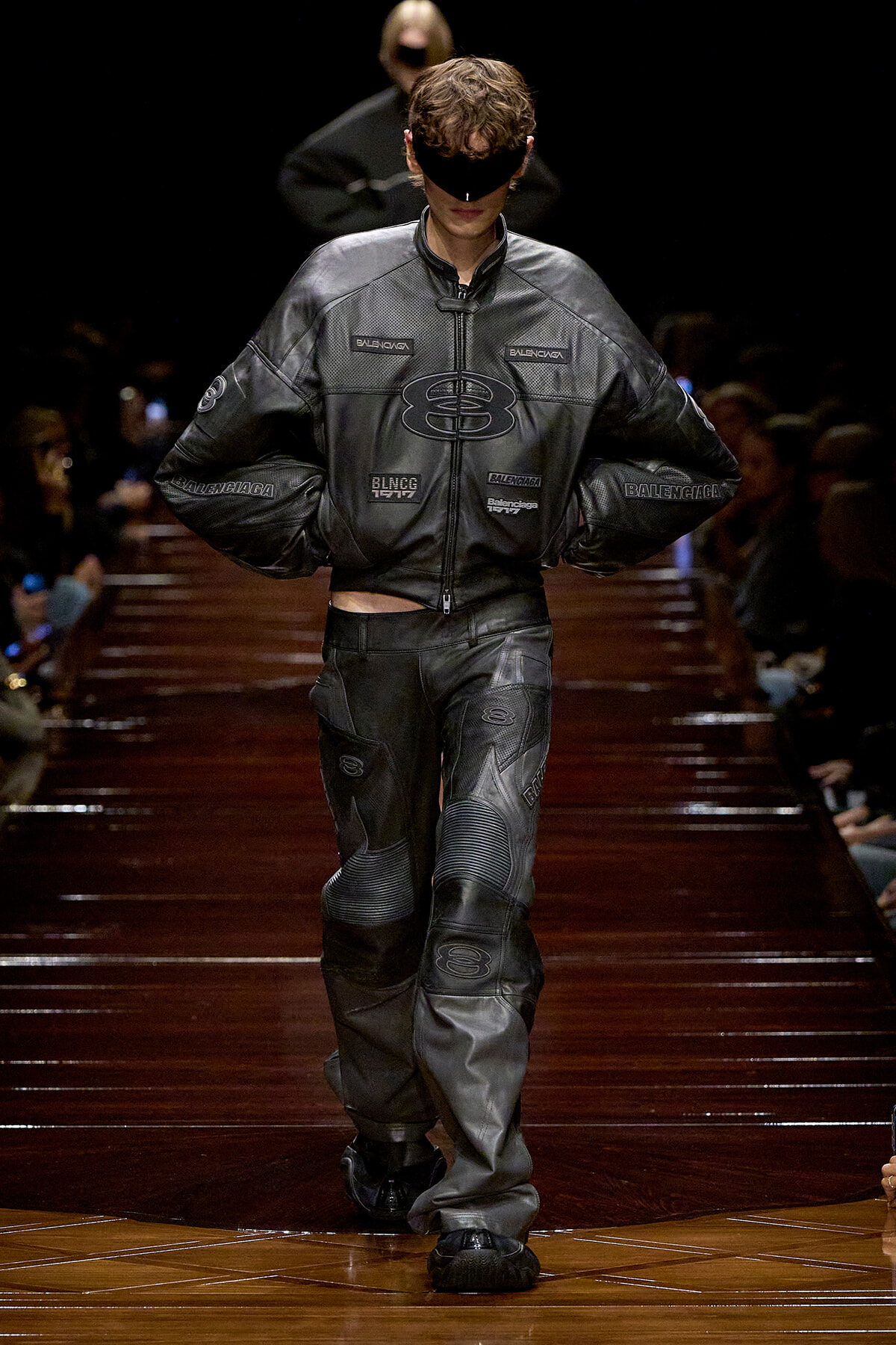 Male model on a dark runway wearing a black leather Balenciaga outfit with a blindfold, hands on hips.