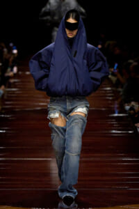 Model on a runway wearing an oversized navy puffer jacket with hood and black sunglasses, paired with ripped denim shorts and jeans.