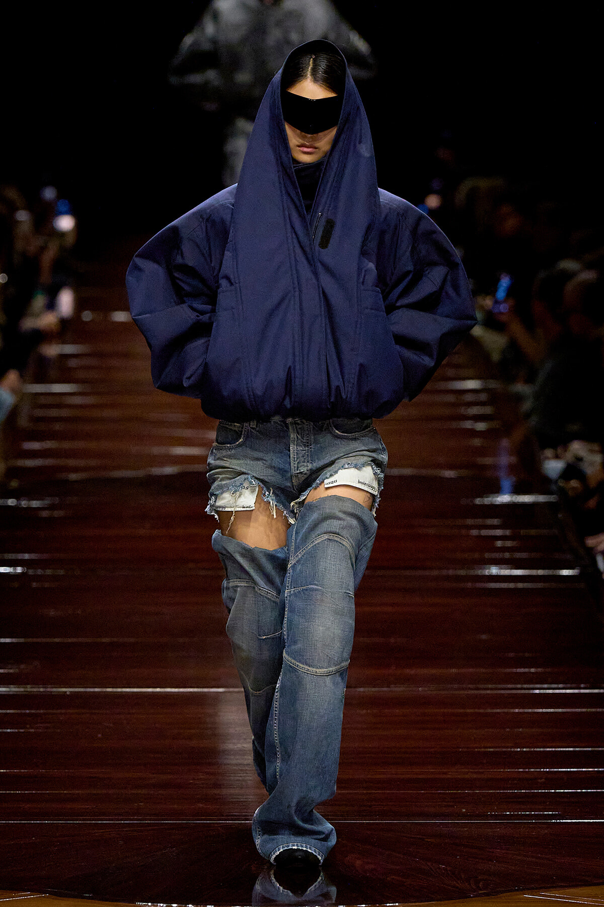 Model on a runway wearing an oversized navy puffer jacket with hood and black sunglasses, paired with ripped denim shorts and jeans.