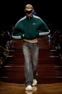 Male model on a dark runway wearing an oversized green zip-up jacket with white striped sleeves, baggy jeans, white sneakers, and a black eye-covering visor; walking forward confidently.
