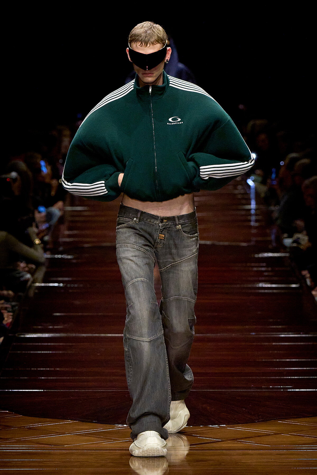 Male model on a dark runway wearing an oversized green zip-up jacket with white striped sleeves, baggy jeans, white sneakers, and a black eye-covering visor; walking forward confidently.