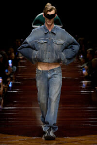 Male model on a runway wearing an oversized cropped denim jacket and matching jeans, with a black visor-style sunglasses piece.