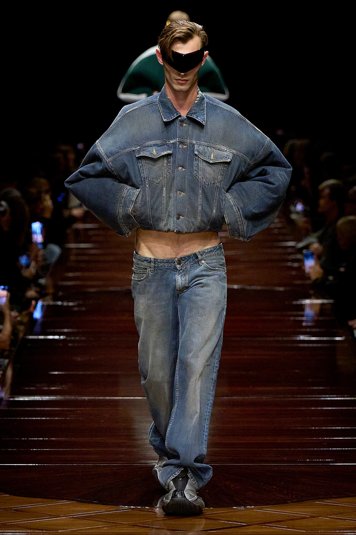 Male model on a runway wearing an oversized cropped denim jacket and matching jeans, with a black visor-style sunglasses piece.