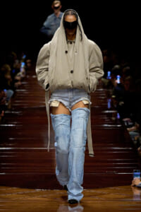 Model walks a runway in an oversized beige trench coat with hood, wearing a black face shield and ripped light-blue jeans; audience in the background.