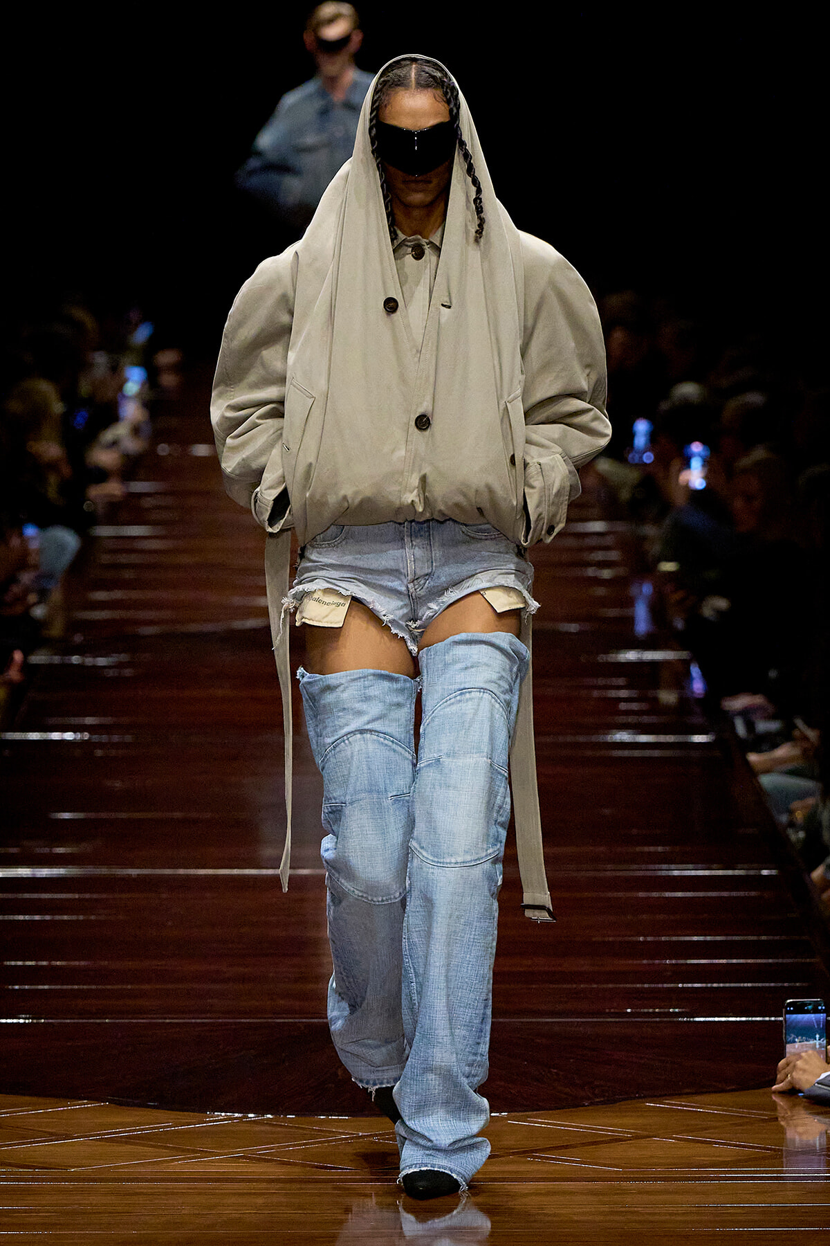 Model walks a runway in an oversized beige trench coat with hood, wearing a black face shield and ripped light-blue jeans; audience in the background.