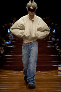 Runway model wearing a beige oversized jacket and patchwork jeans, with a black futuristic visor over the eyes.