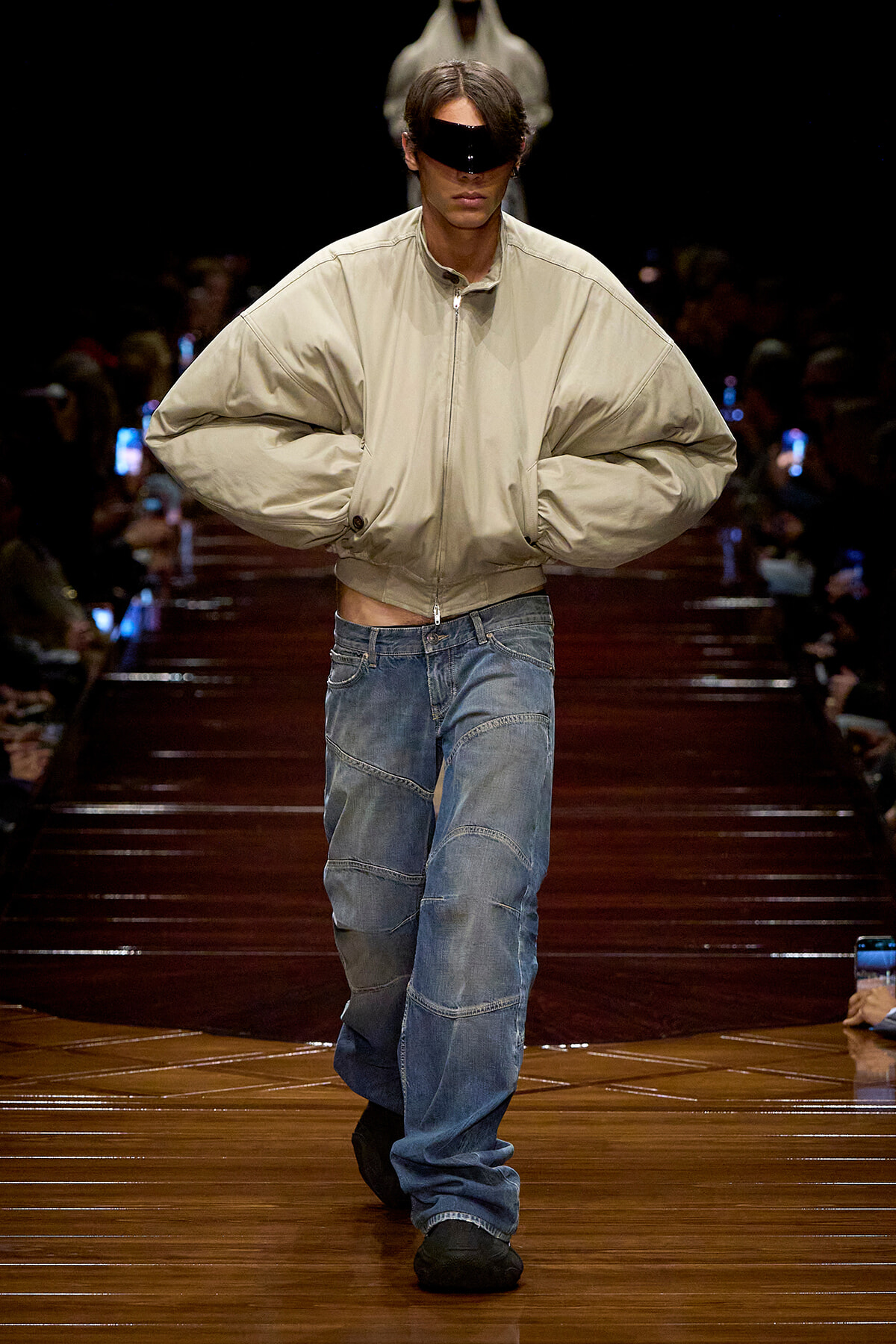Runway model wearing a beige oversized jacket and patchwork jeans, with a black futuristic visor over the eyes.
