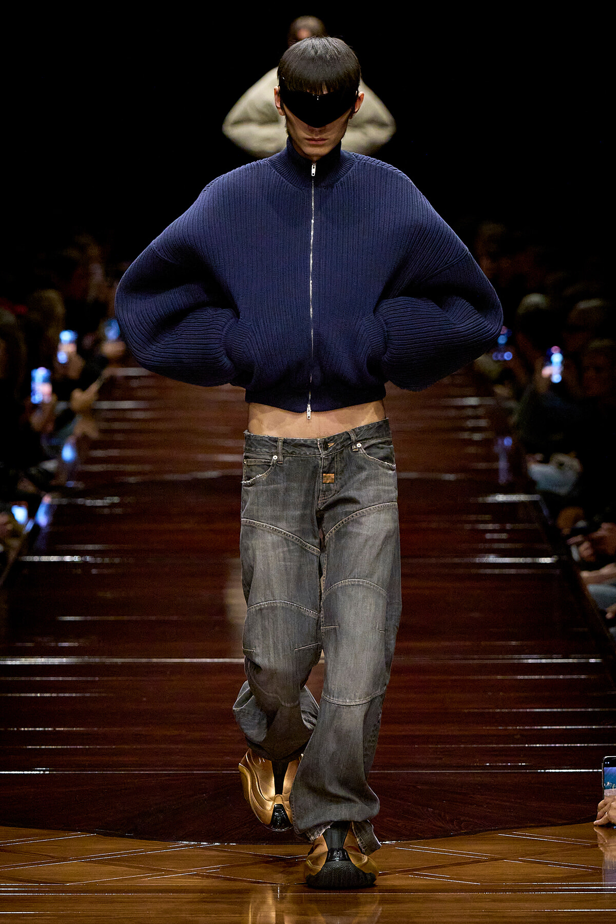 Model walks the runway in an oversized navy knit zip-up sweater and baggy denim jeans, wearing futuristic dark visor sunglasses.