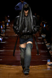 Model walks the runway in a voluminous black leather hooded coat with a wide belt and oversized sleeves, paired with distressed denim thigh-high boots.