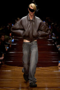 Model walks a runway in an oversized brown leather jacket with exaggerated sleeves and loose gray jeans, wearing a black eye-covering visor.