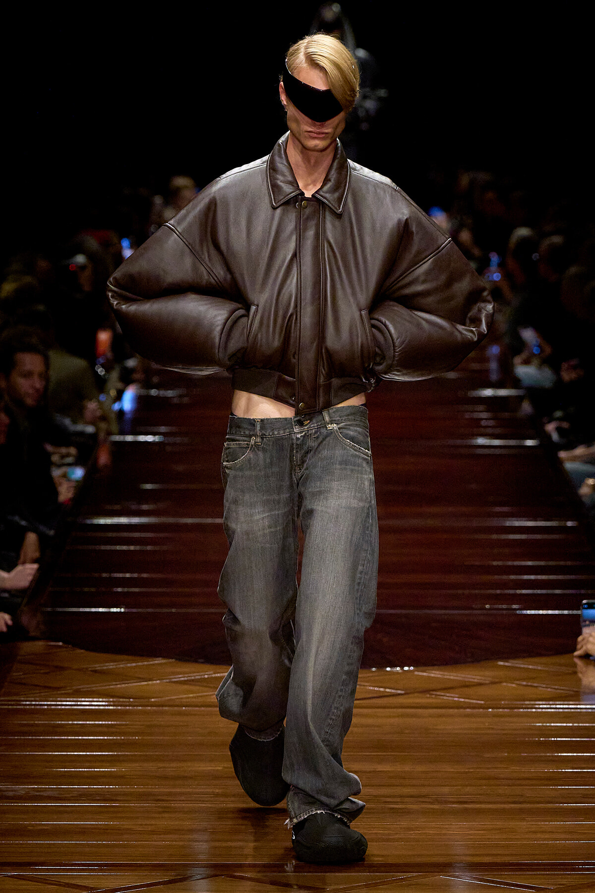Model walks a runway in an oversized brown leather jacket with exaggerated sleeves and loose gray jeans, wearing a black eye-covering visor.