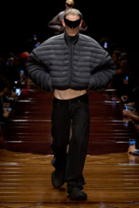 Model walks a runway in an oversized black puffer jacket, cropped at the midriff, paired with black wide-leg pants and a futuristic visor.
