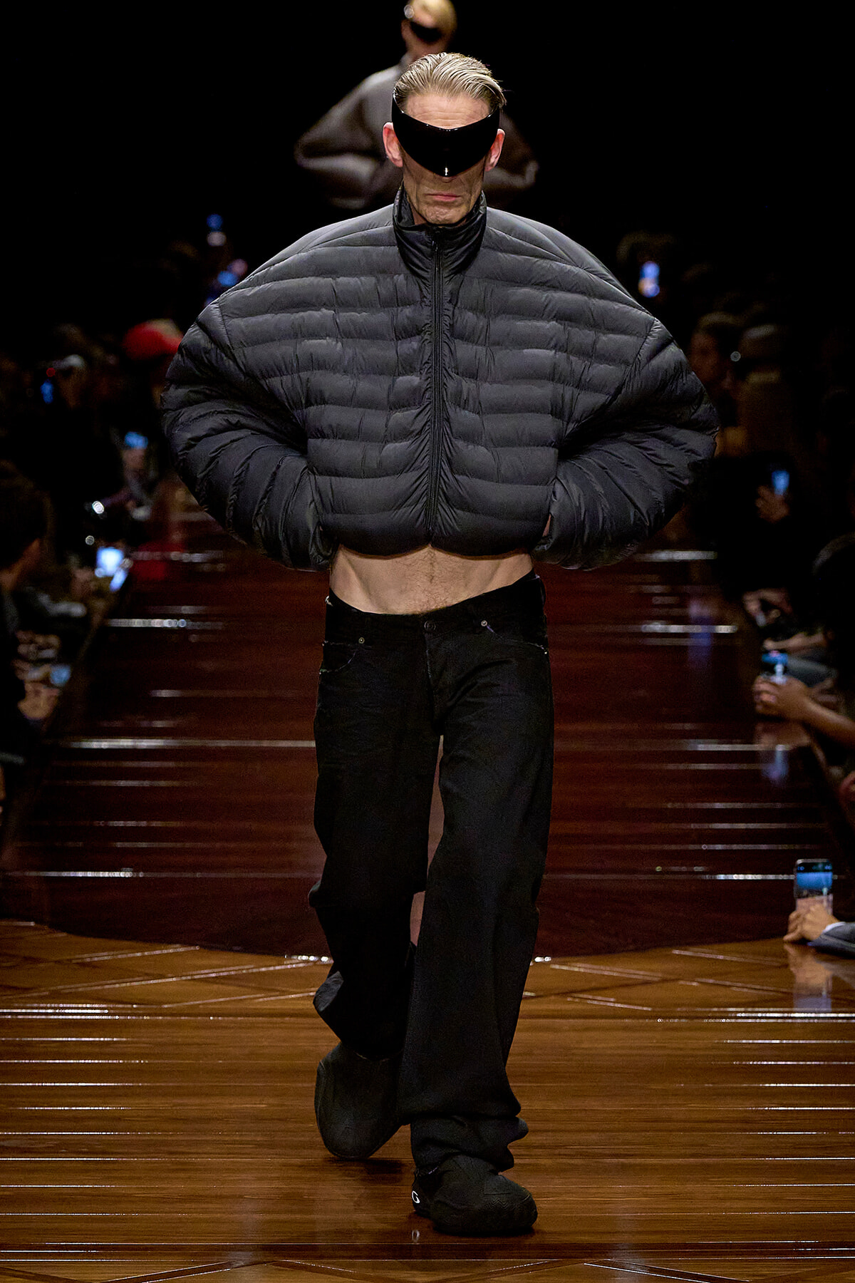 Model walks a runway in an oversized black puffer jacket, cropped at the midriff, paired with black wide-leg pants and a futuristic visor.