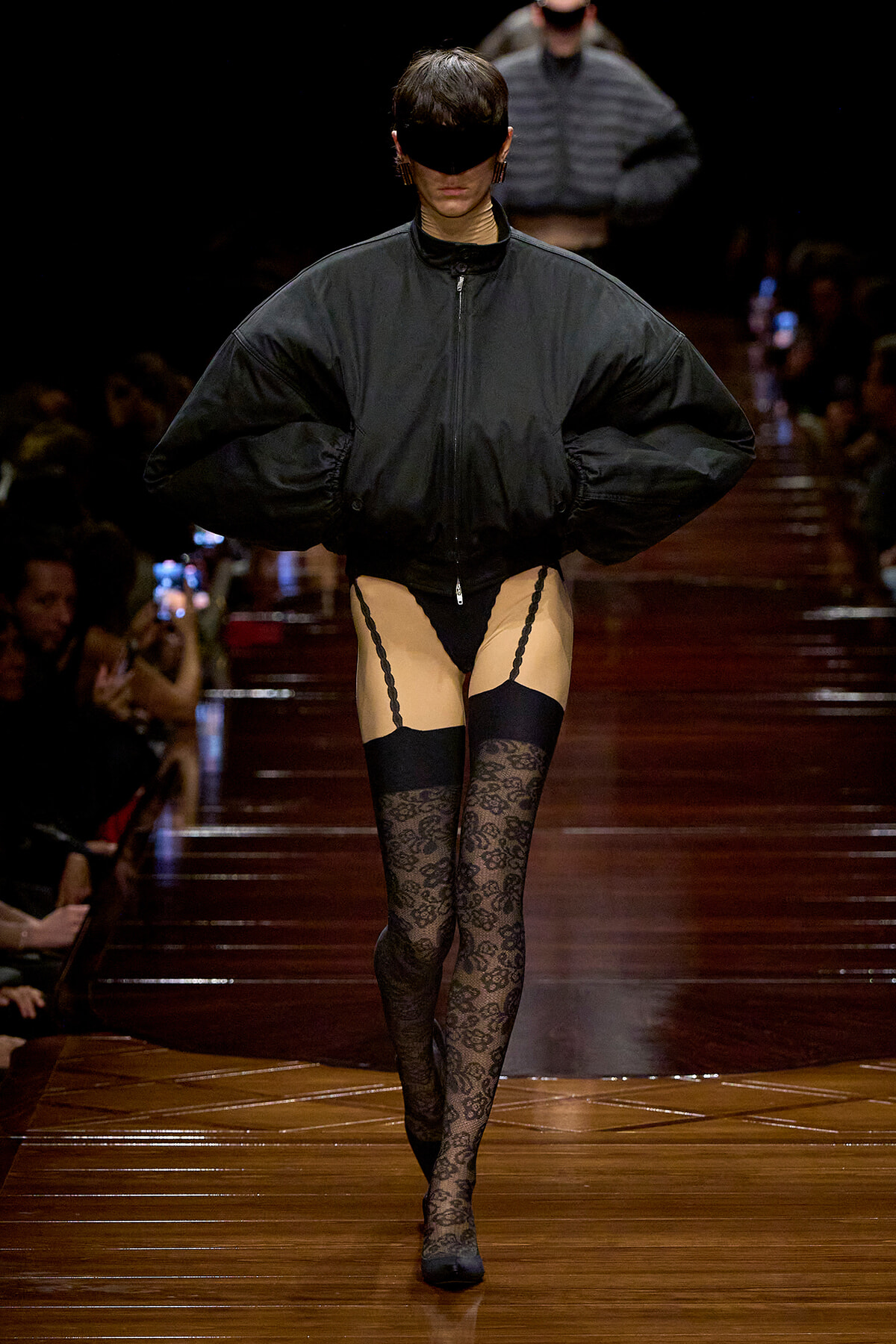 Runway model wearing an oversized black jacket, black eye mask, and sheer lingerie with lace-thigh stockings in a fashion show.