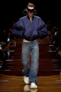 Male runway model in an oversized navy bomber jacket with baggy patchwork jeans and white sneakers walks the catwalk with a black visor over his eyes.
