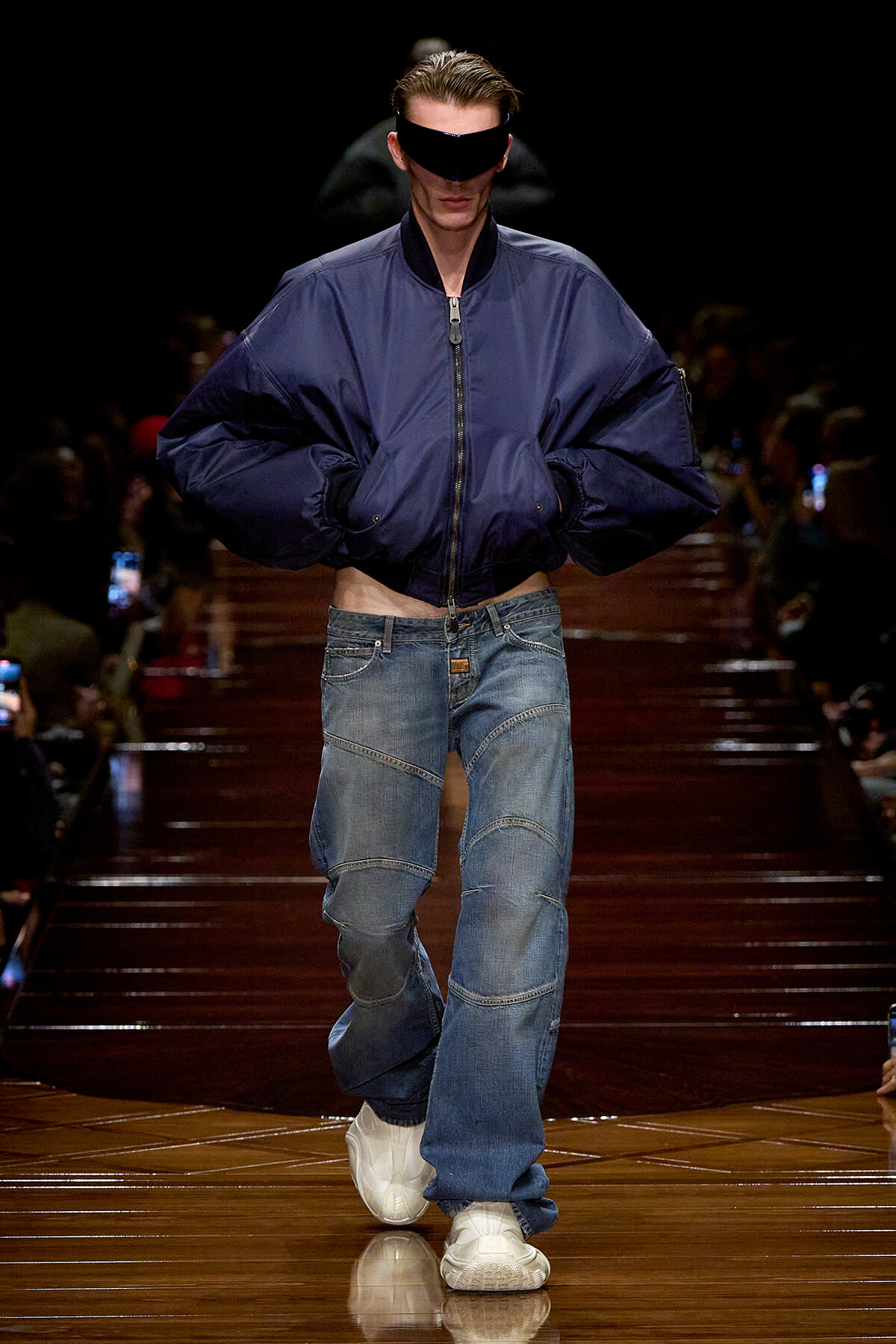 Male runway model in an oversized navy bomber jacket with baggy patchwork jeans and white sneakers walks the catwalk with a black visor over his eyes.