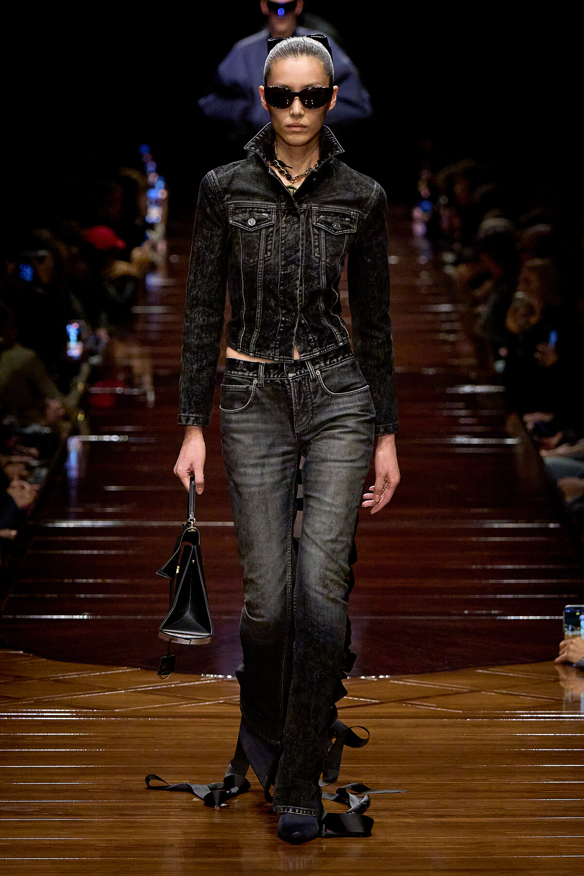 Model walks a dark denim-on-denim outfit on a runway, wearing sunglasses and carrying a black handbag.