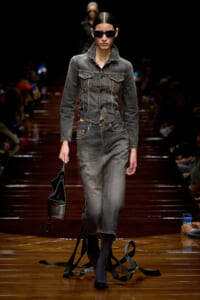 Model on a runway wearing a gray denim jacket and matching jeans, with sunglasses and a black handbag in hand.