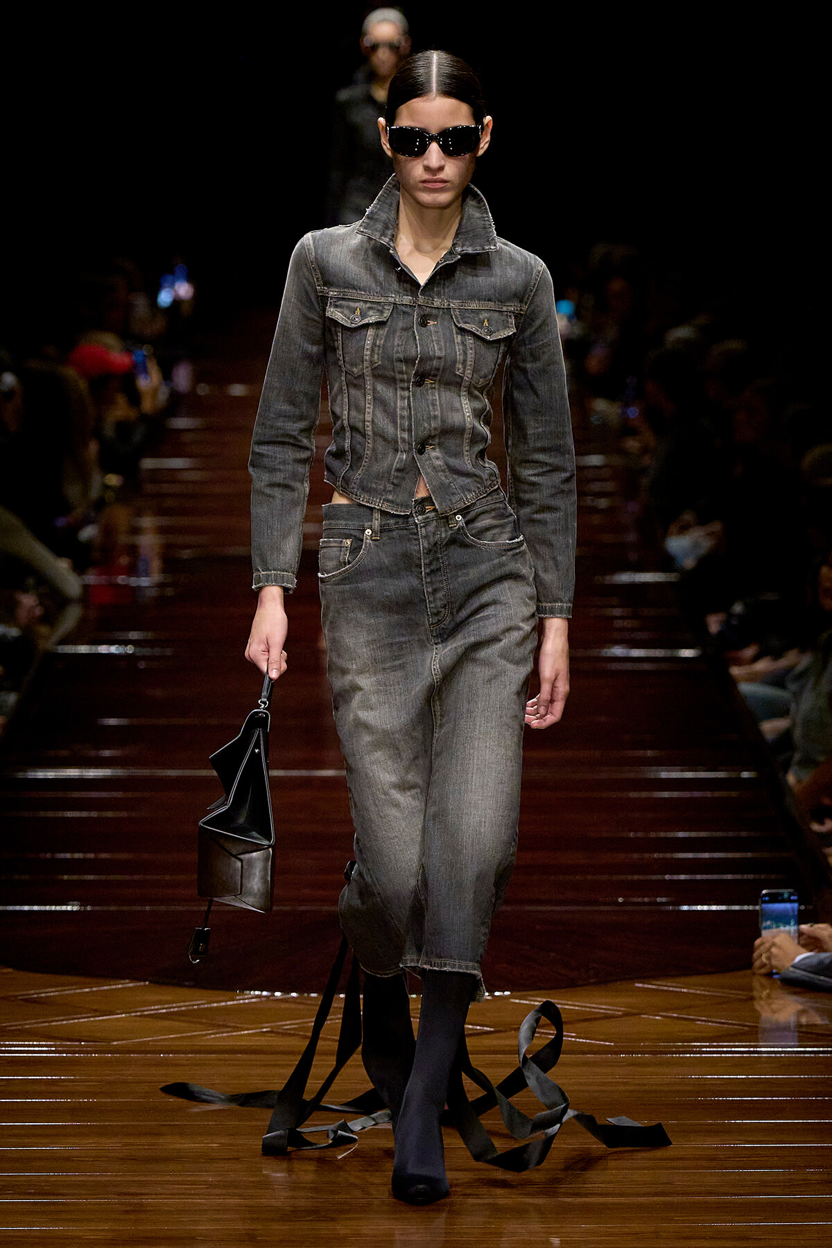 Model on a runway wearing a gray denim jacket and matching jeans, with sunglasses and a black handbag in hand.