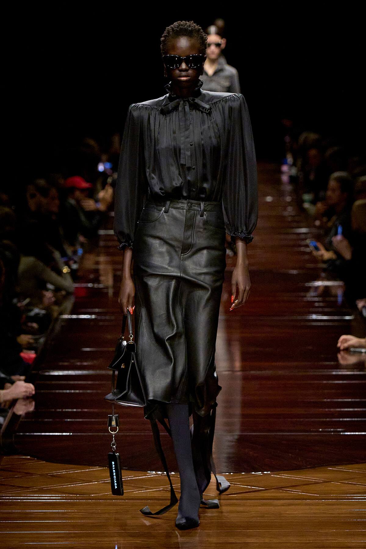 Model walks the runway in an all-black ensemble: satin blouse with voluminous sleeves, leather midi skirt, oversized sunglasses, and a small black handbag on her arm.