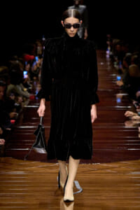 Model walking a dark velvet midi dress with high neck and puffed sleeves on a glossy runway, wearing sunglasses and carrying a black handbag.