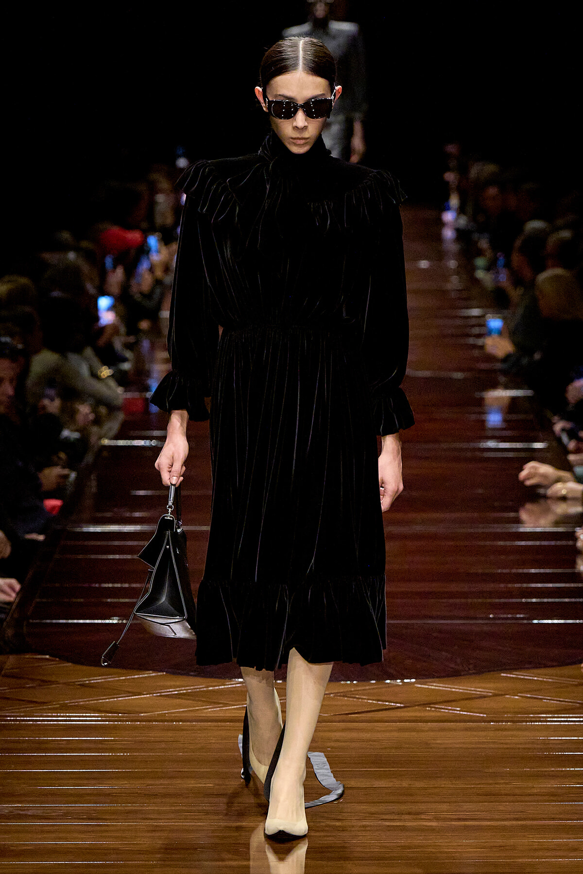 Model walking a dark velvet midi dress with high neck and puffed sleeves on a glossy runway, wearing sunglasses and carrying a black handbag.