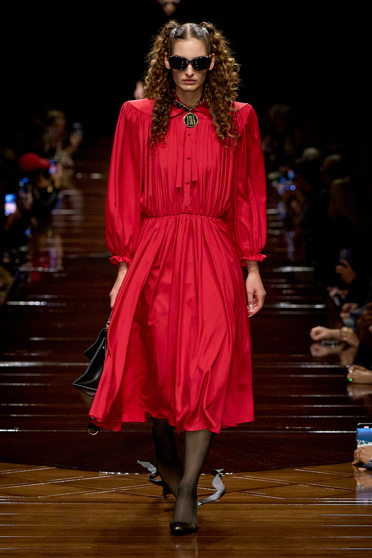 Model walks the runway in a bright red midi dress with long sleeves and a gathered waist, wearing dark sunglasses and carrying a black handbag.