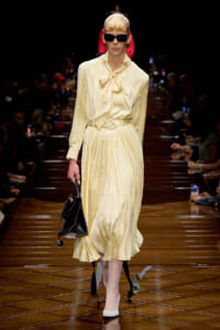 Model walks the runway in a pale yellow satin blouse with a bow and a pleated midi skirt, belted, wearing dark sunglasses and carrying a black handbag.