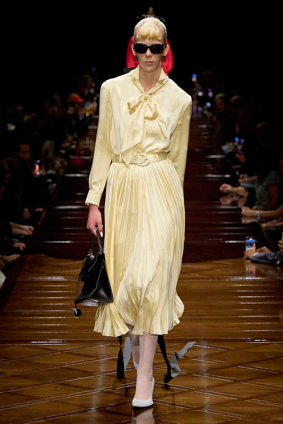 Model walks the runway in a pale yellow satin blouse with a bow and a pleated midi skirt, belted, wearing dark sunglasses and carrying a black handbag.