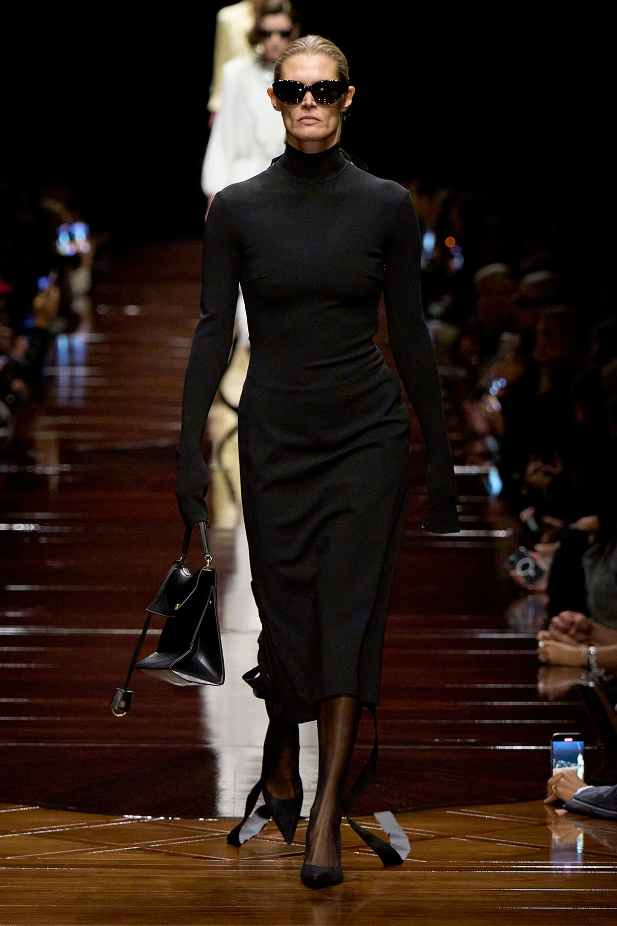 Model walking a runway in a black turtleneck midi dress, oversized sunglasses, and black heels, carrying a structured black handbag.
