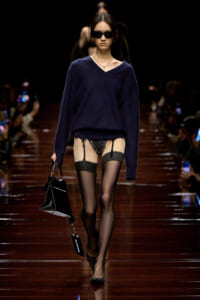 Model walking a runway in a navy oversized sweater, sheer black stockings with a garter belt, black heels, sunglasses, and a black handbag.