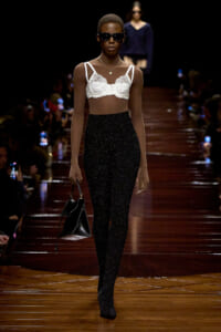 Model walks a fashion runway in a white lace bralette, glittery black high-waisted pants, black sunglasses, and a small black handbag.