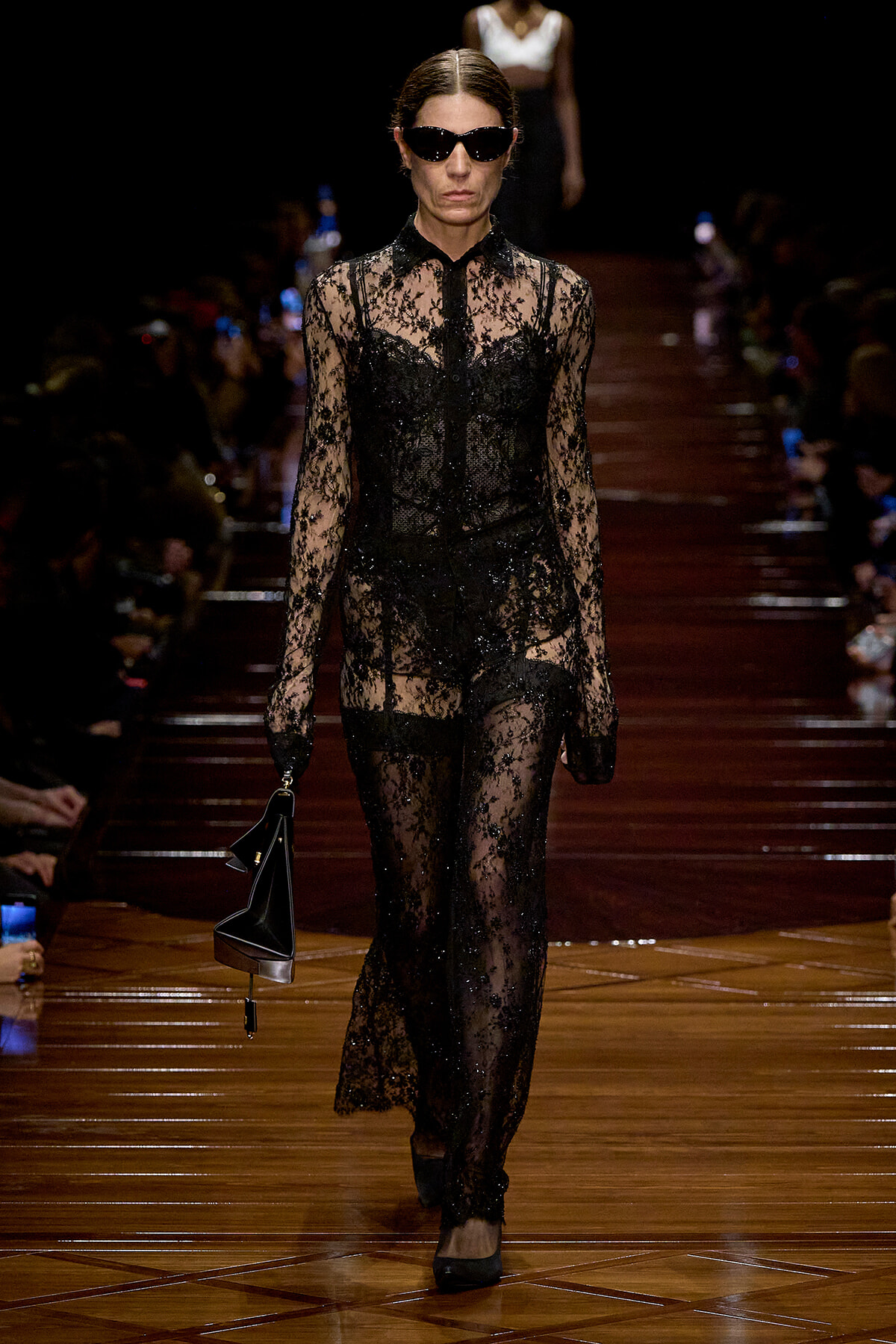 Model on a runway wearing a full-length black lace sheer gown with gloves and sunglasses, carrying a black handbag.