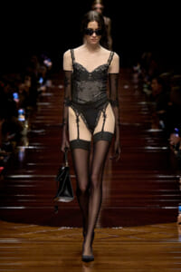 Model walks a black lace lingerie bodysuit with sheer panels, thigh-high stockings, and garter straps on a wooden runway. She wears dark sunglasses and carries a small black handbag.
