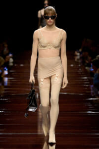 Model walking a runway in beige lace lingerie set with thigh-high stockings, sunglasses, and a small black handbag.