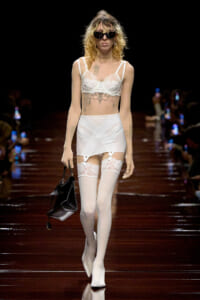 Model walking a runway in white lace lingerie with garter belt and thigh-high stockings, sunglasses, and a black handbag.