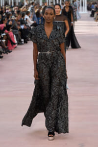 Model walking a runway in a floor-length black dress with silver geometric patterns and a thin belt.