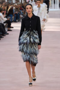Model walking a runway in a black, textured coat with a dramatic feathered blue-gray skirt and metallic heels.