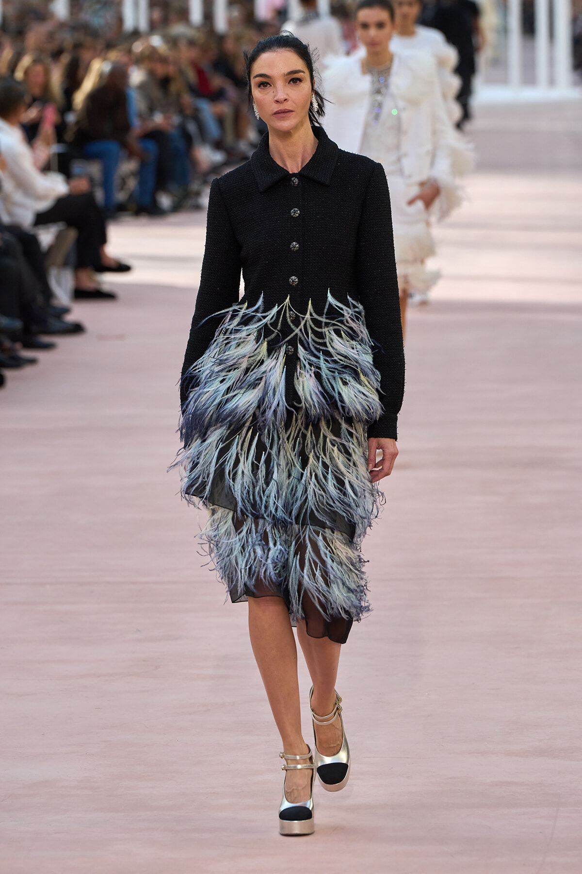 Model walking a runway in a black, textured coat with a dramatic feathered blue-gray skirt and metallic heels.
