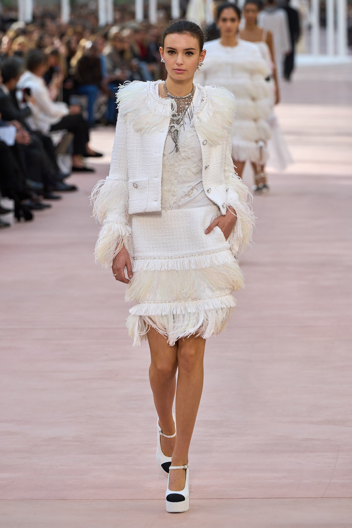 Model walking the runway in a white textured suit with feathered fringe and heavy jewelry.