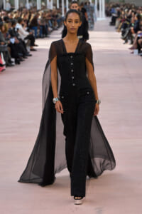 Model walking a fashion runway in a black sequined jumpsuit with a sheer cape flowing behind her, audience on both sides.