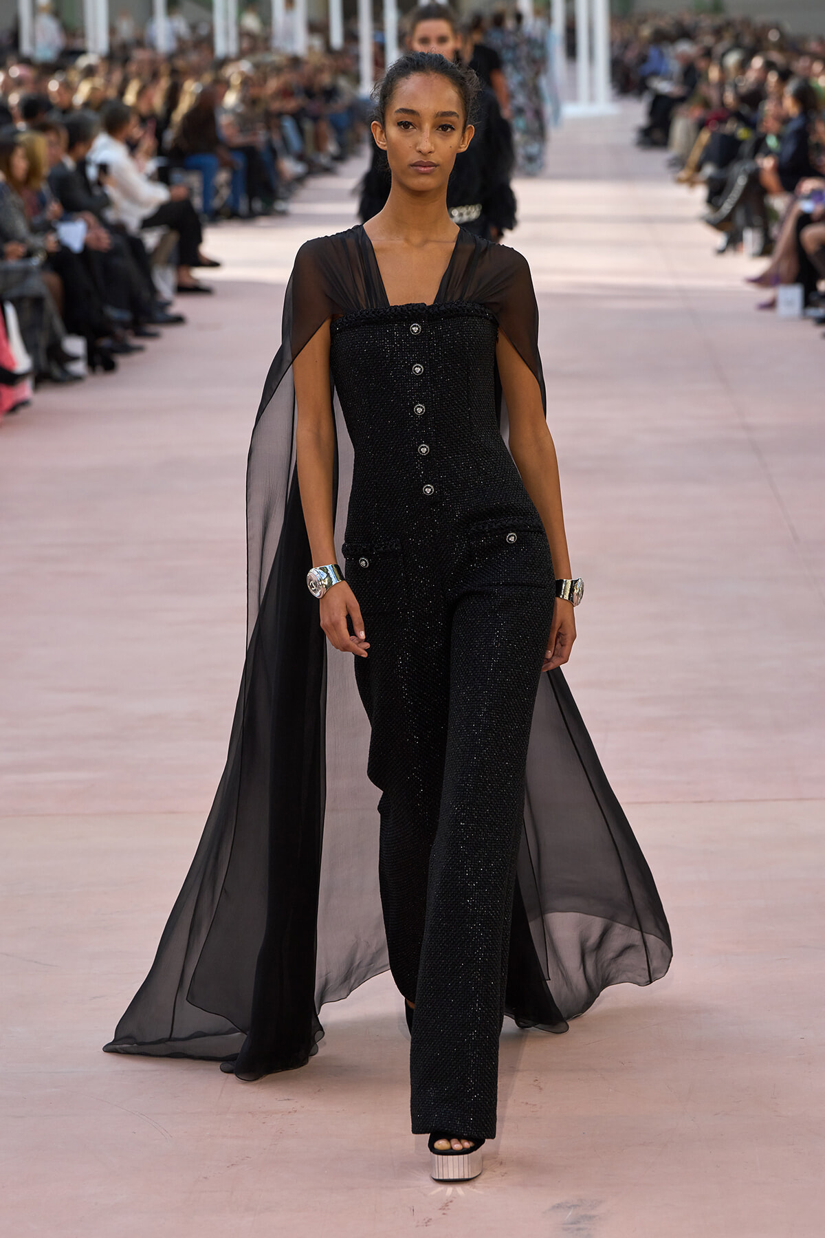 Model walking a fashion runway in a black sequined jumpsuit with a sheer cape flowing behind her, audience on both sides.