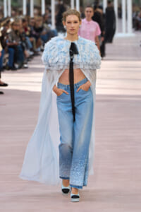 Model walks a runway wearing a pale blue feathered cape with black ribbon, light-wash jeans with glitter at the cuffs, and blue platforms.