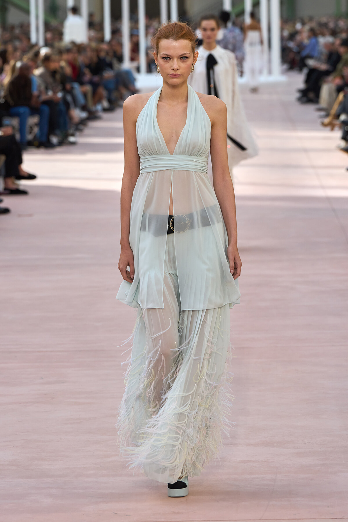 Model walks a pastel blue halter gown with a sheer overlay and feathered fringe along the hem on a fashion runway